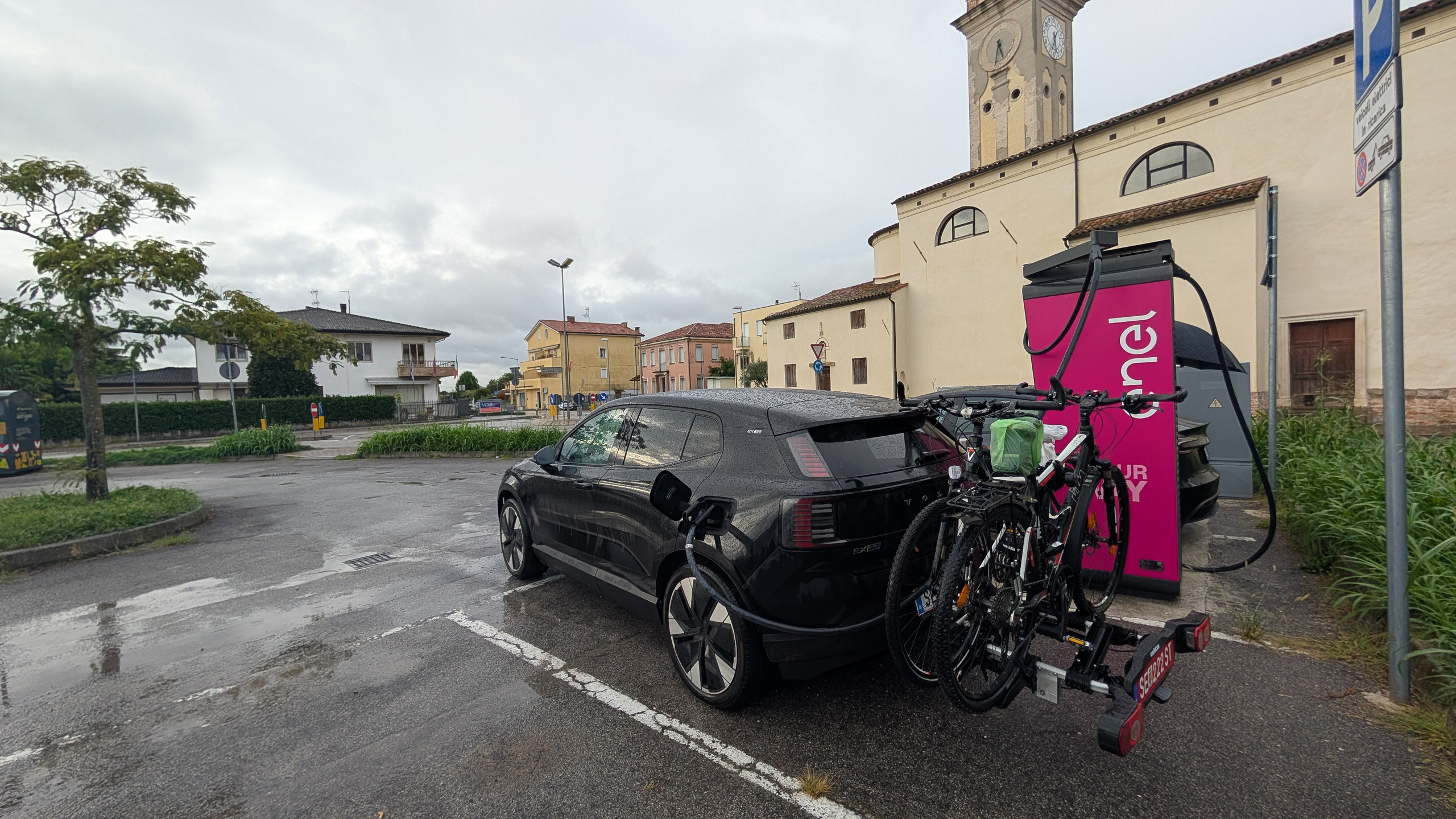 Ladestopp im Regen – E-Auto mit Rädern am Heckträger vor einer Dorfkirche